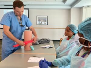 A man in medical scrubs bandages a model leg while two teens dressed in medical scrubs look on
