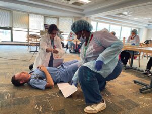 Two people, one dressed in a doctor’s white coat and another in disposable PPE, kneeling over another person laying flat on the ground. They are securing a long piece of cloth, a pelvic binder, around the person on the ground. In the background, there are other people in PPE watching the demonstration.